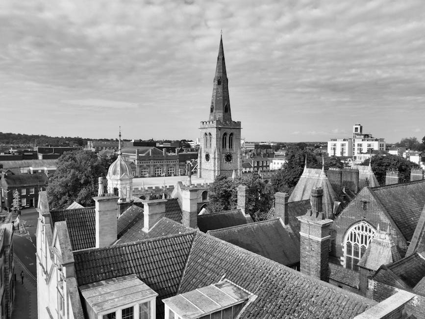 Black and white photograph showing a view over rooftops in Balham, with a prominent church steeple featuring arched windows and a clock, surrounded by chimneys, dormer windows, and building rooftops made of tiles and brick. In the background, there are modern buildings and trees under a partly cloudy sky. The image is taken from an elevated position, highlighting the dense arrangement of residential and historic structures typical of a home relocation setting. There are no visible objects related to packing or moving equipment in this scene, but the shot reflects a typical neighborhood in Balham, SW12, where furniture transport and house removals by Removal Van Balham could occur during a relocation process.