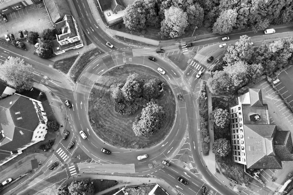An aerial black-and-white photograph showing a roundabout in an urban residential area with multiple cars navigating the circular traffic flow. In the center of the roundabout, there is a small, landscaped island featuring three large trees and grass. Surrounding the roundabout are multi-story buildings, including houses and a larger apartment complex, with trees lining the streets and parking areas filled with parked cars. Vehicles are seen entering and exiting the roundabout, which is part of a home relocation process or furniture transport. The scene illustrates street-level movement and the coordination involved in loading or unloading furniture and boxes during a removal service. This visual detail aligns with house removals and moving logistics, highlighting the environment where Removal Van Balham might facilitate the effective transit of household items, documented via a detailed aerial view that captures the spatial arrangement, road markings, and vehicle positions, emphasizing the traffic flow during a furniture transfer or home packing and moving procedure.
